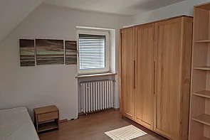 Zimmer des Hufeland-Hauses für Freiwillige, mit Fenster, Bett, Kleiderschrank, einem offenen Schrank und einem Nachttisch.